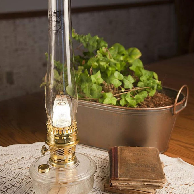 Clear glass oil lamp with brass burner glowing softly on a lace doily beside worn vintage books and a potted green ivy plant, ideal for Lamps & Lighting, Parts and Accessories, and Lamp and Lantern Fuel collections.