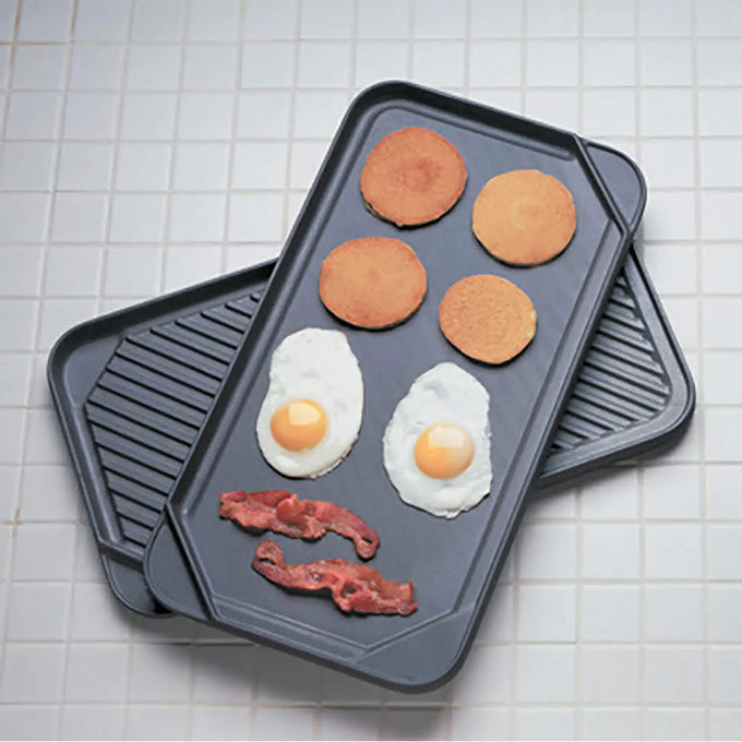 Two black reversible pans stacked on a white tiled surface with pancakes, fried eggs, and strips of bacon cooking on the smooth griddle side, featured in Kitchen & Food, Cookware, Pots and Pans, USA Made, Best Sellers, and Promo Eligible collections.