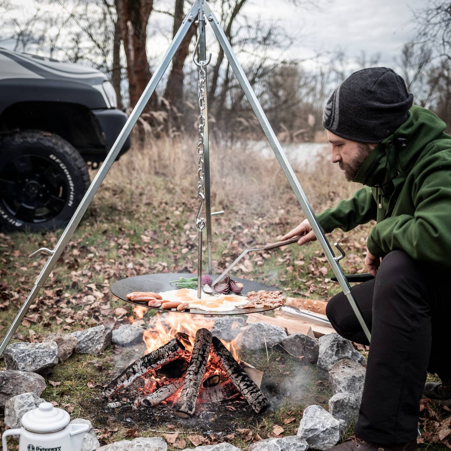 Man cooking breakfast outdoors using a sturdy metal tripod with adjustable chain suspension over a campfire, ideal for Farm, Garden & Tool, Camping, Best Sellers, and Promo Eligible collections.