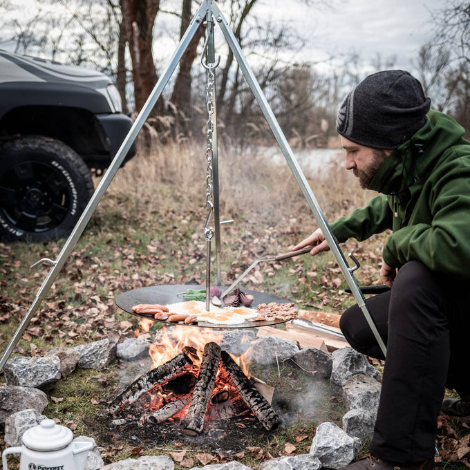 Man cooking breakfast outdoors using a sturdy metal tripod with adjustable chain suspension over a campfire, ideal for Farm, Garden & Tool, Camping, Best Sellers, and Promo Eligible collections.