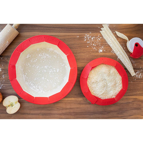 Two red silicone pie crust shields, one fitted over uncooked dough and the other around a baked pie, displayed on a wooden surface with baking tools and apple slices nearby, ideal for Kitchen & Food and Baking Supplies collections.