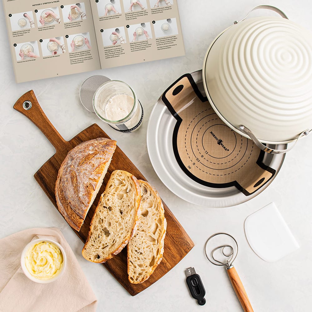 Overhead view of freshly sliced sourdough bread on a wooden board next to a white ceramic baking cloche, dough scraper, whisk, measuring cup, and open recipe booklet in a Kitchen & Food setting.