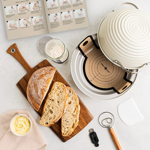 Overhead view of freshly sliced sourdough bread on a wooden board next to a white ceramic baking cloche, dough scraper, whisk, measuring cup, and open recipe booklet in a Kitchen & Food setting.
