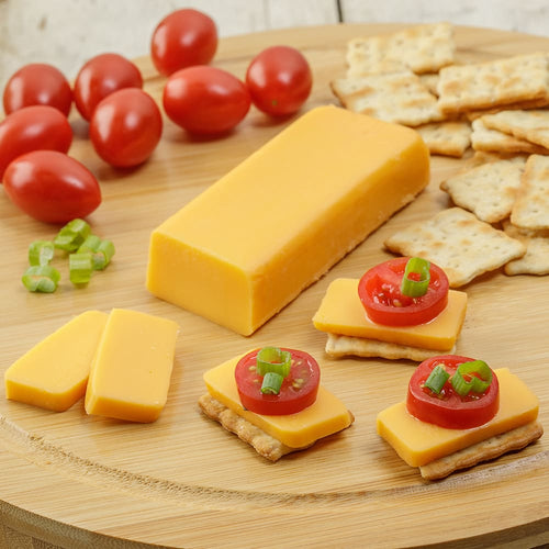 Block of smooth cheddar cheese with sliced pieces arranged on a wooden board alongside crackers, grape tomatoes, and chopped green onions, highlighting a shelf-stable cheese option for Kitchen & Food, Pantry, and USA Made collections.