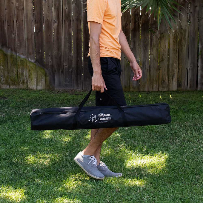 Person carrying a black carrying case for handcrafted ladder toss game on a grassy lawn with wooden fence background, suitable for Toys, Books & Games and Lawn Games collections.