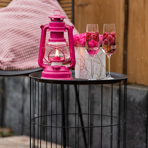 Handcrafted bright pink galvanized oil lantern with thermal heat-resistant globe glowing warmly on a black wireframe side table next to two raspberry-filled champagne flutes and pink roses, ideal for Lamps & Lighting, Camping, and Emergency Supplies collections.