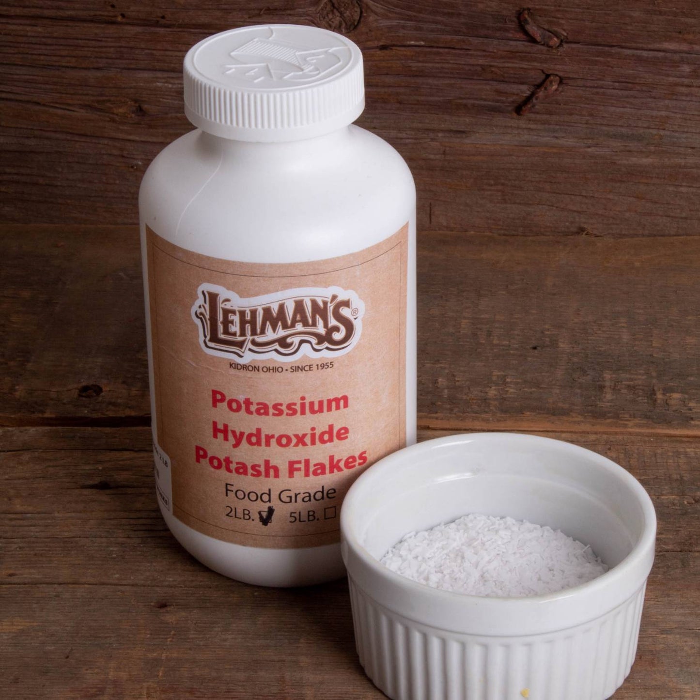 White plastic container of food-grade potassium hydroxide potash flakes beside a small white ceramic bowl filled with white flakes on a wooden surface, featured in soapmaking and health collections.