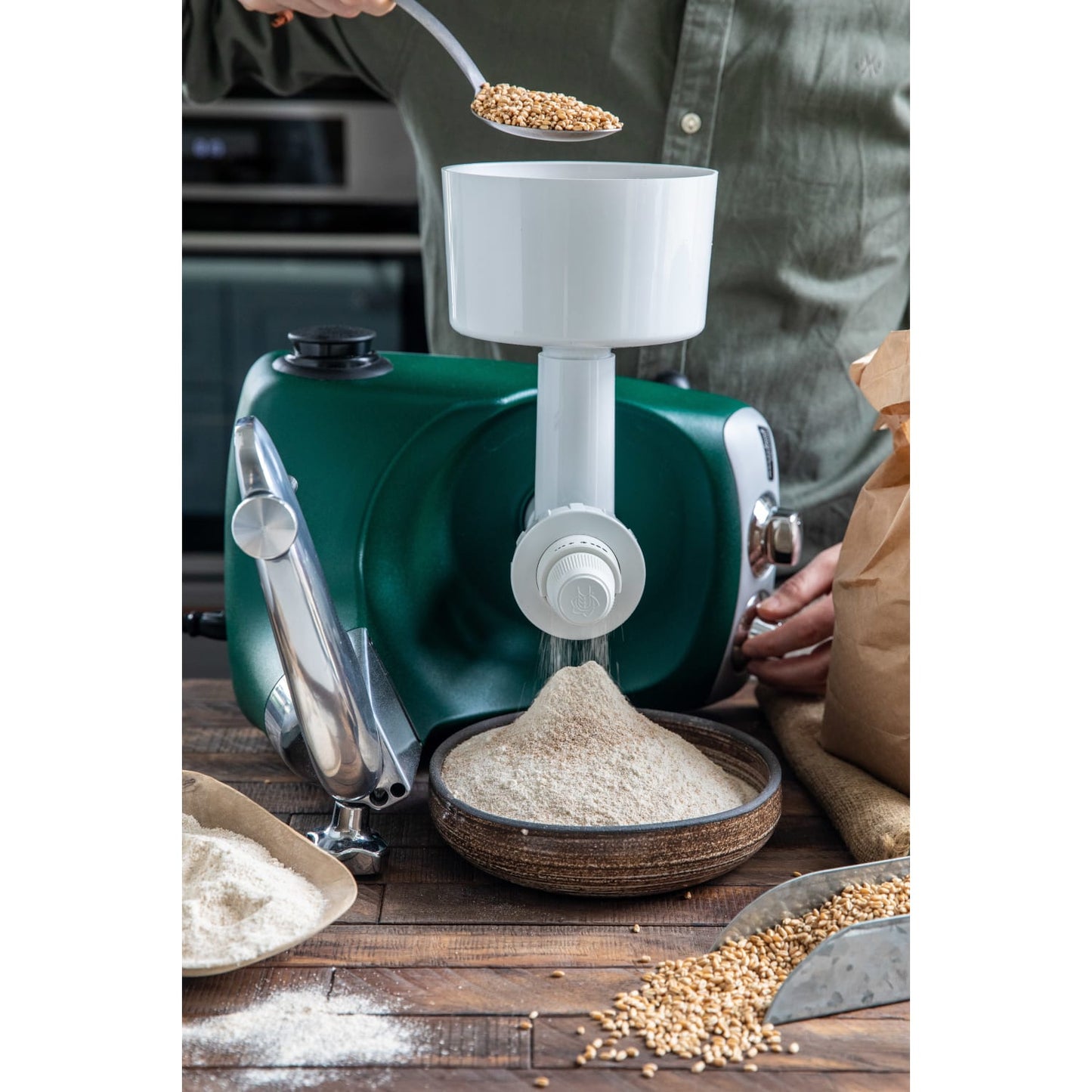 Green stand mixer fitted with a white grain mill attachment grinding fresh flour into a bowl on a wooden surface with scattered wheat grains and bowls, ideal for Kitchen & Food, Baking Supplies, Grain Mills, Bread Making Supplies, and Kitchen Gadgets collections.