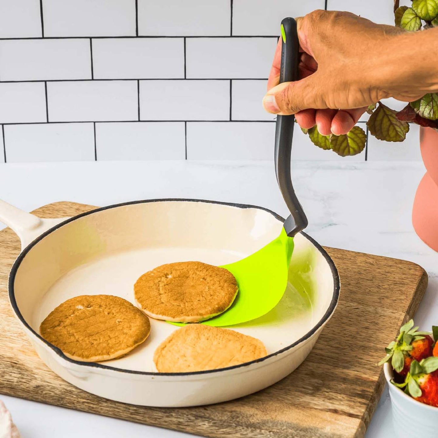 Hand holding a flexible spatula with a bright green blade and black handle lifting a pancake in a white enamel skillet on a wooden board, ideal for Kitchen & Food and Kitchen Gadgets collections.