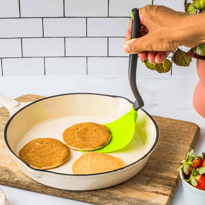 Hand holding a flexible spatula with a bright green blade and black handle lifting a pancake in a white enamel skillet on a wooden board, ideal for Kitchen & Food and Kitchen Gadgets collections.