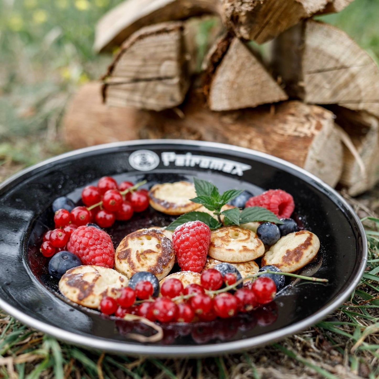 Black enamel plate from Petromax holding golden brown potato pancakes with fresh red currants, raspberries, blueberries, and mint leaves, set outdoors by stacked firewood, perfect for Farm, Garden & Tool, Kitchen & Food, Camping, Dinnerware, Best Sellers, and Promo Eligible collections.