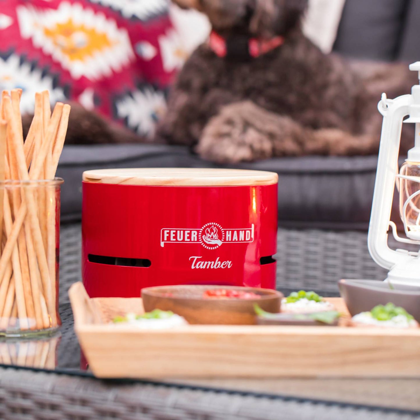 Bright red Feuerhand Tamber Tabletop Charcoal Grill with a wooden lid on a glass outdoor table among snacks and a lantern, featured in Farm, Garden & Tool and Camping collections.