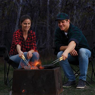 Two people using a handcrafted cast iron double pie iron with wooden handles over an open fire, enjoying outdoor cooking in a forest setting, perfect for camping and farm collections.
