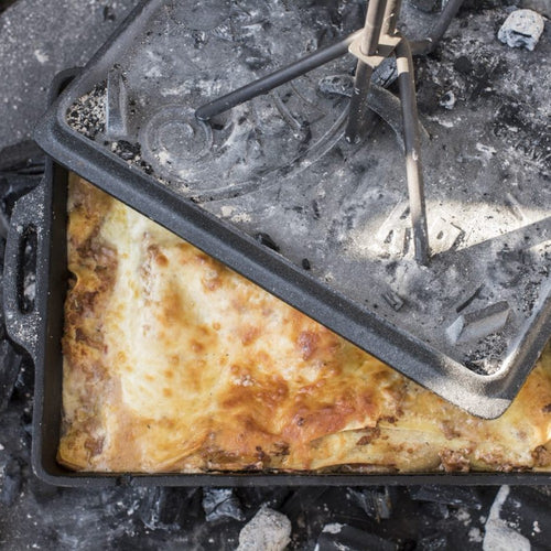 Cast iron baking pan with a golden-brown baked dish inside, set on hot ash and embers, illustrating outdoor cooking in the Farm, Garden & Tool and Camping collections.