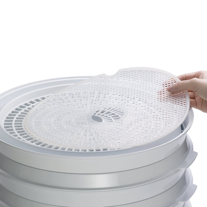 Flexible nonstick mesh screen being placed on stack of white dehydrator trays, designed to prevent small foods from falling through, ideal for food preservation and kitchen use