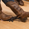 Black painted cast iron boot jack shaped like a cricket, assisting removal of brown leather boots on a hardwood floor, part of Home & Decor and Household Helpers best sellers.