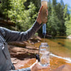 Person outdoors using a compact water filter to purify murky river water into a clear portable bottle, ideal for camping, emergency supplies, and outdoor adventures.