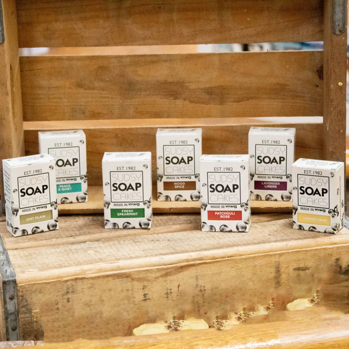 Seven uniquely scented handmade bar soaps in eco-friendly packaging arranged on a rustic wooden shelf, highlighting natural ingredients and handcrafted USA made wellness products.