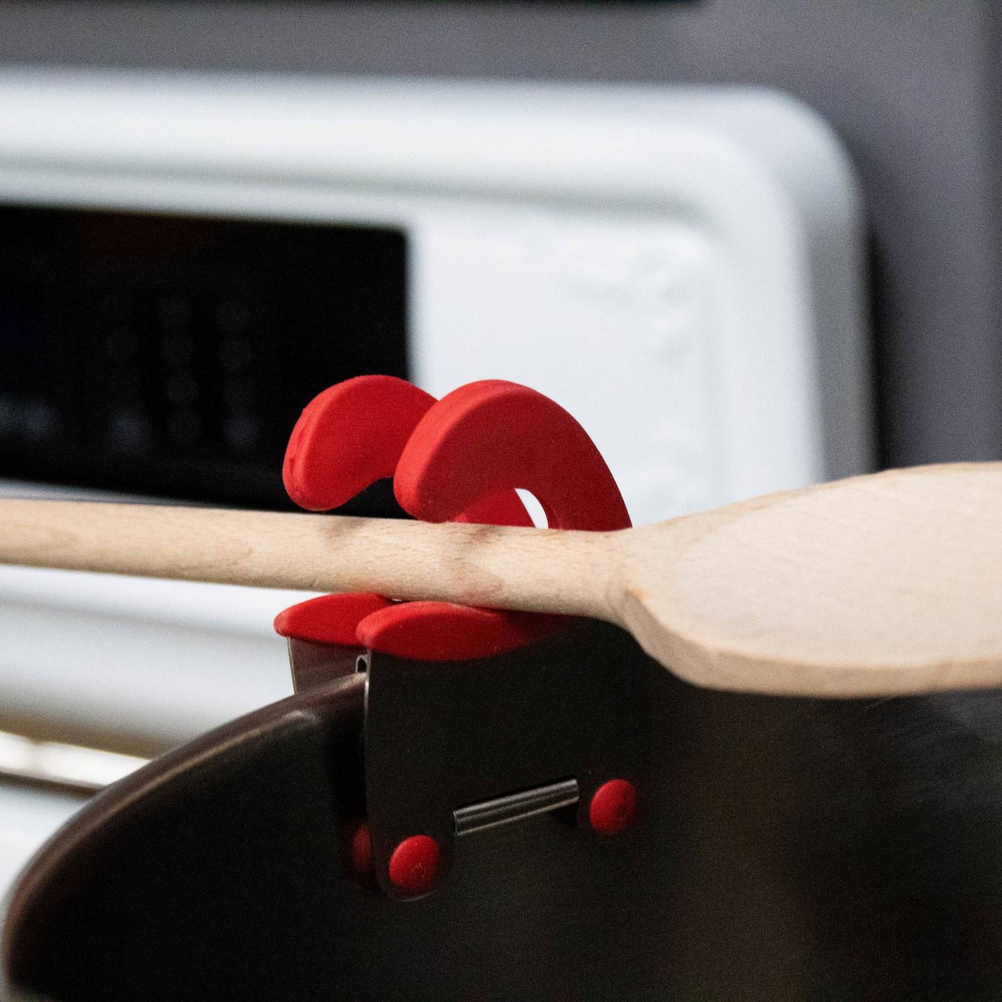 Close-up of a pot clip spoon holder with red silicone grips clamped to a black pot holding a wooden spoon, designed to keep utensils off surfaces and within easy reach in Kitchen & Food and Kitchen Gadgets collections.