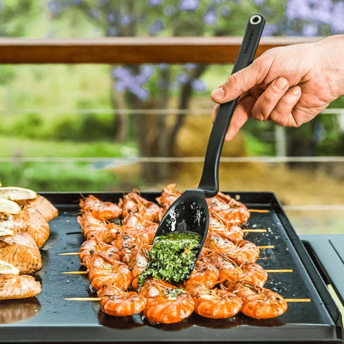 Hand holding a black basting brush with a deep, flexible scooping head spreading green sauce over shrimp skewers on a flat top grill, ideal for Kitchen & Food and Grilling collections.