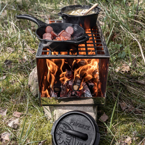 Compact handcrafted metal camp stove with active wood fire burning inside, cooking sausage in cast iron skillet and stew in pot, ideal for Farm, Garden & Tool, Camping, Emergency Supplies, and Best Sellers collections.