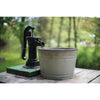 Black vintage hand water pump mounted on a green wooden base next to a weathered gray metal bucket, set on a rustic wooden table outdoors with a blurred natural background, featured in Home and Garden How-To Books and Best Sellers collections.