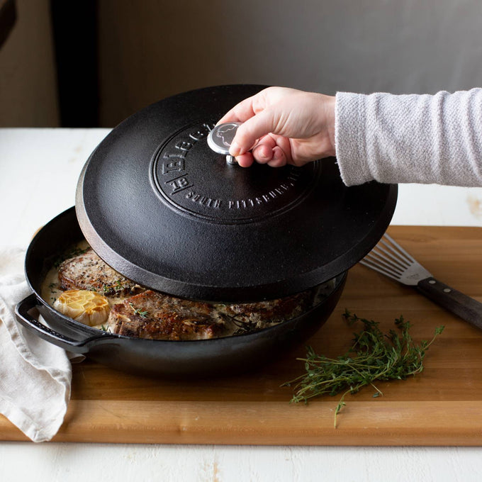 Hand lifting a triple-seasoned black cast iron lid from skillet with cooked food inside, resting on wooden surface with fresh herbs, ideal for kitchen and cookware collections.