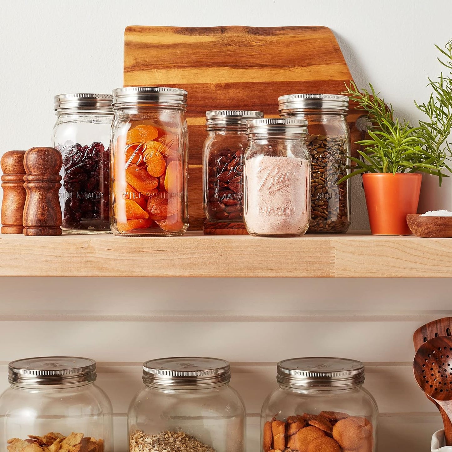 Three wide-mouth jars with sleek stainless steel lids arranged on a kitchen shelf, surrounded by dried fruits, spices, wooden salt and pepper shakers, a wooden cutting board, and a small potted plant, showcasing elegant storage in Kitchen & Food and Storage and Serving collections.