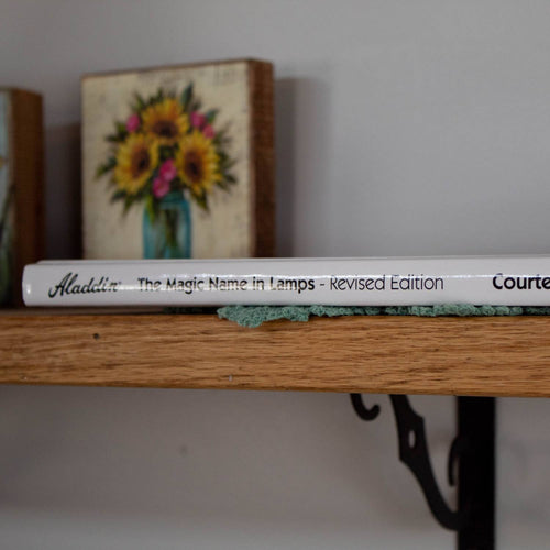 Side view of a handcrafted hardcover book titled "The Magic Name in Lamps - Revised Edition" resting on a wooden shelf, with a small decorative painting of sunflowers in a blue vase in the background, ideal for Toys, Books & Games, Heritage and Lifestyle Books, and Best Sellers collections.