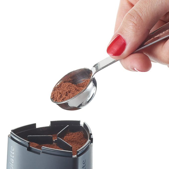 Hand holding a small metal scoop with cocoa powder above a black circular stencil holder with six compartments for creating latte art designs, featured in Brewing Coffee and Tea, Kitchen Gadgets, and Best Sellers collections.