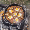 Large cast iron skillet cooking breakfast hash with diced potatoes, vegetables, and five sunny-side-up eggs over an open outdoor fire, featured in Kitchen & Food, Cookware, Cast Iron, USA Made, and Best Sellers collections.