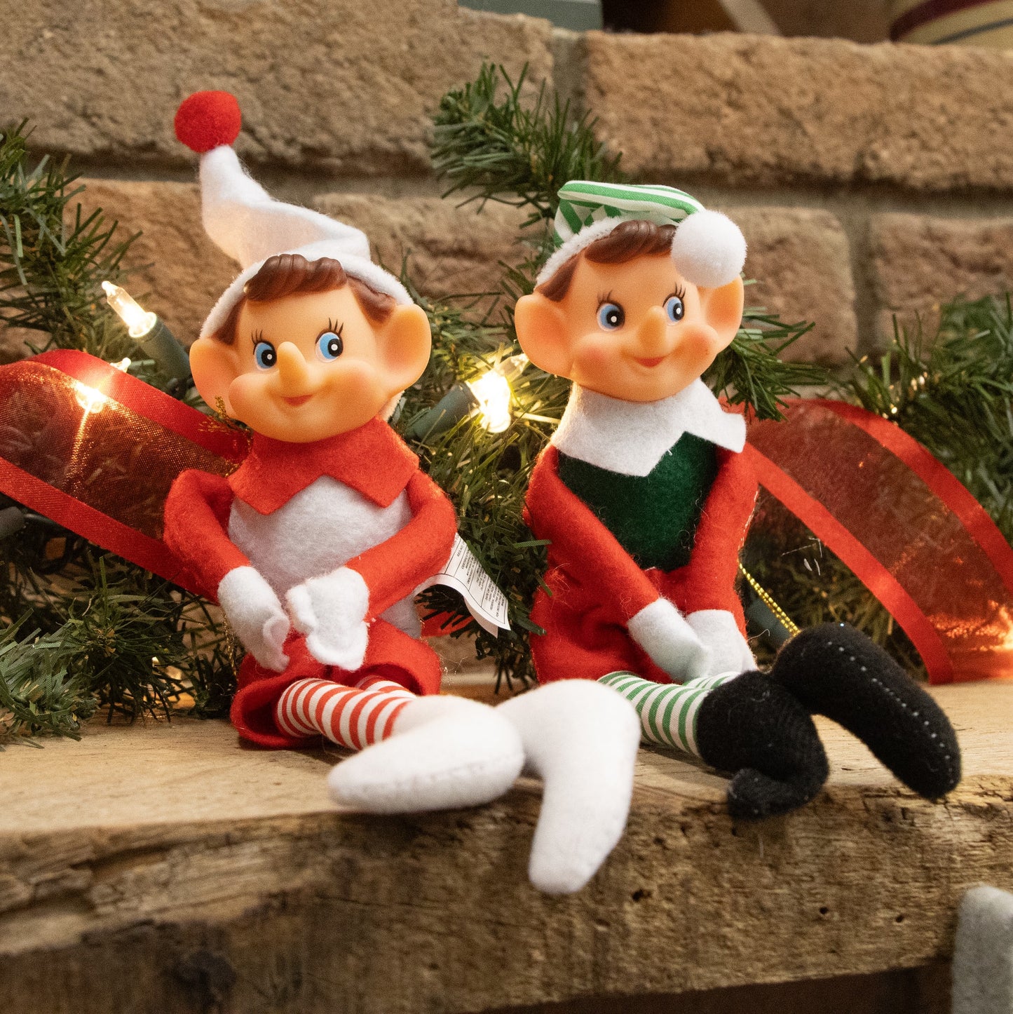 Two posable fabric pixie elves in festive red, green, and white outfits with striped stockings and felt hats, seated on a wooden surface with holiday decor in Home & Decor and Holiday Decor collections.