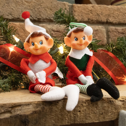 Two posable fabric pixie elves in festive red, green, and white outfits with striped stockings and felt hats, seated on a wooden surface with holiday decor in Home & Decor and Holiday Decor collections.