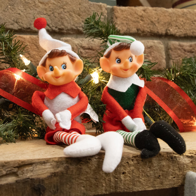 Two posable fabric pixie elves in festive red, green, and white outfits with striped stockings and felt hats, seated on a wooden surface with holiday decor in Home & Decor and Holiday Decor collections.