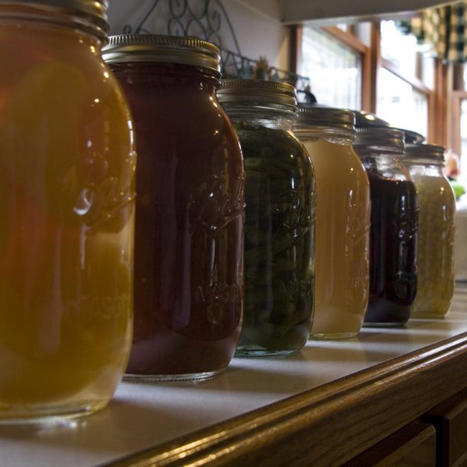 Row of clear glass regular-mouth jars filled with various preserved foods, displayed on a kitchen counter near a sunlit window, fitting Kitchen & Food, Food Preservation, Canning, USA Made, and Best Sellers collections.