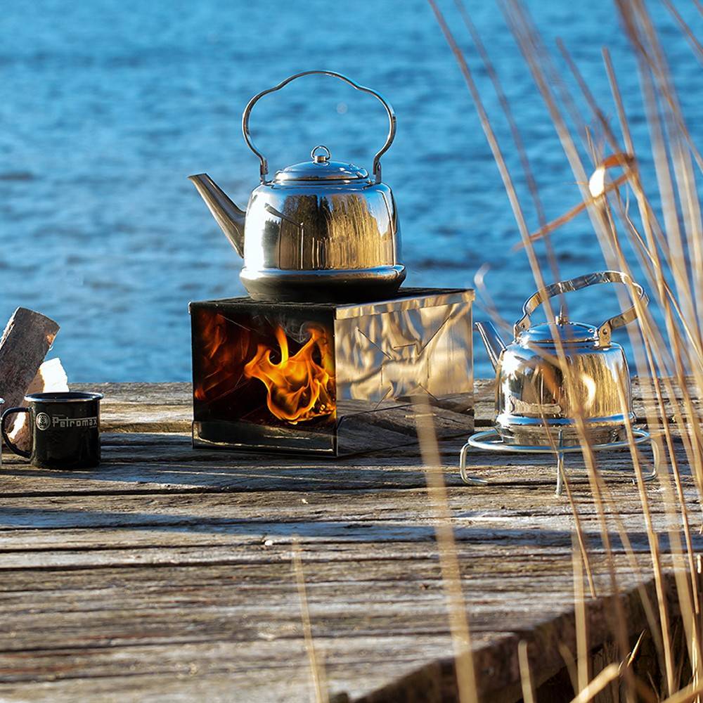 Polished stainless steel teakettle boiling water over an open flame on a portable stove by a lakeside, alongside a second kettle and a metal camping mug, suited for Farm, Garden & Tool, Kitchen & Food, Camping, and Brewing Coffee and Tea collections.
