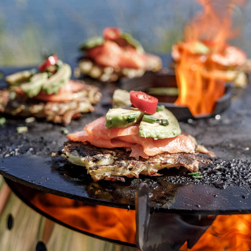 Close-up of juicy grilled salmon topped with avocado slices, chives, and a red chili pepper on a handcrafted steel griddle with open flame, ideal for outdoor cooking in camping and grilling collections.