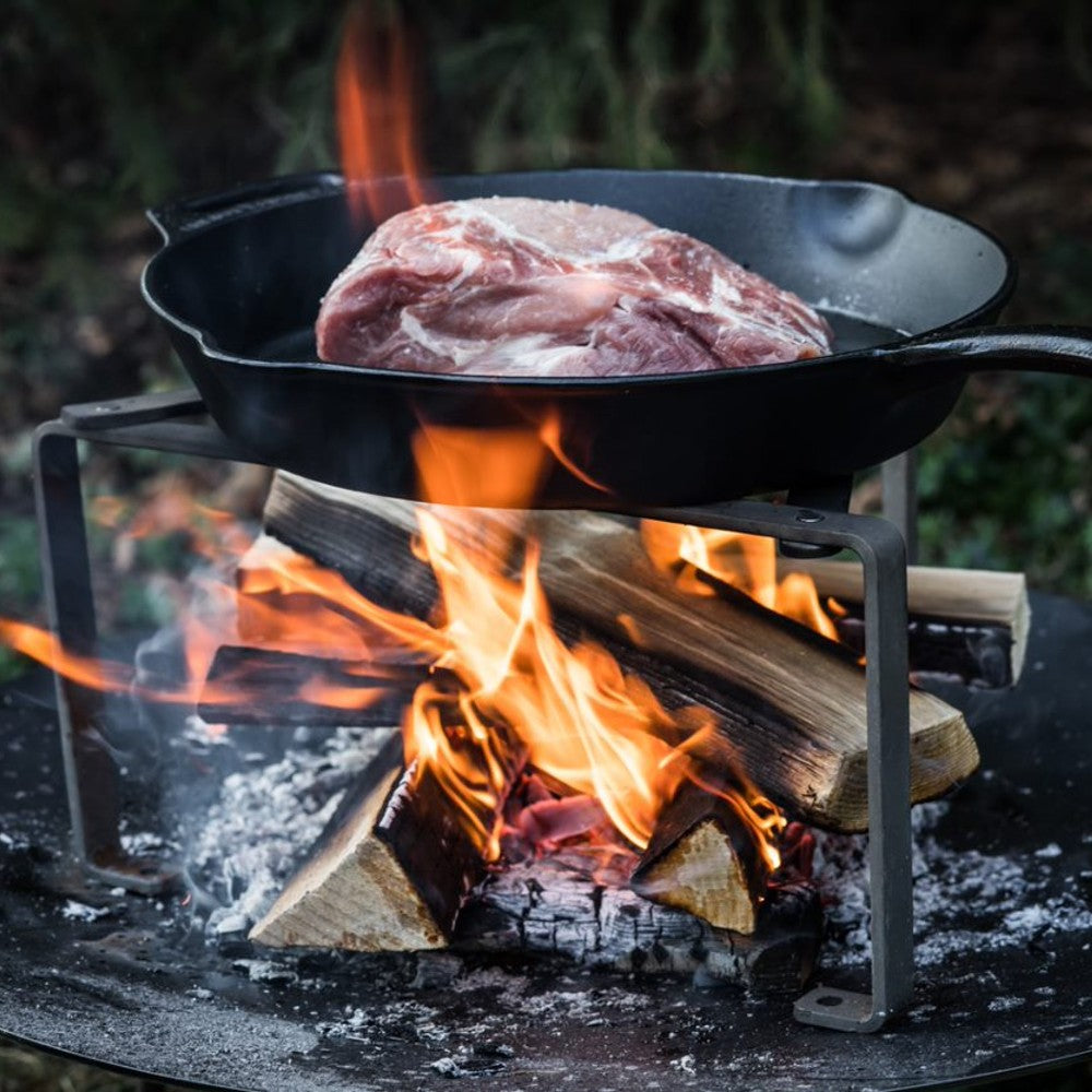 Heavy-duty steel stand supporting a cast iron skillet with raw meat over an open wood fire, ideal for outdoor cooking in Farm, Garden & Tool, Camping, and Best Sellers collections.