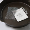Handcrafted stainless steel chainmail scrubber resting inside a cast iron skillet beside a white drawstring pouch with vintage-style logo, perfect for Farm, Garden & Tool and Kitchen & Food collections.