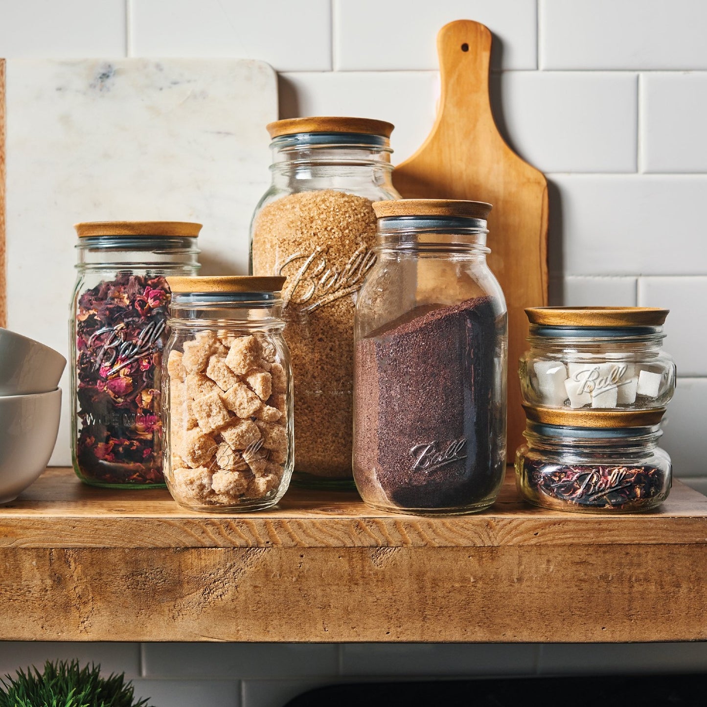 Set of six clear glass kitchen jars with Ball branding, topped with airtight acacia wood lids, filled with various pantry staples on a rustic wooden shelf, styled for Kitchen & Food and Storage and Serving collections.
