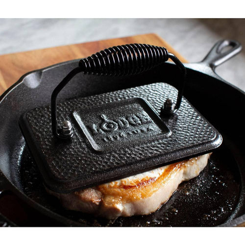 Seasoned cast iron rectangular grill press with coiled handle resting on a searing pork chop in a skillet, perfect for Kitchen & Food, Cast Iron, and Cookware collections.