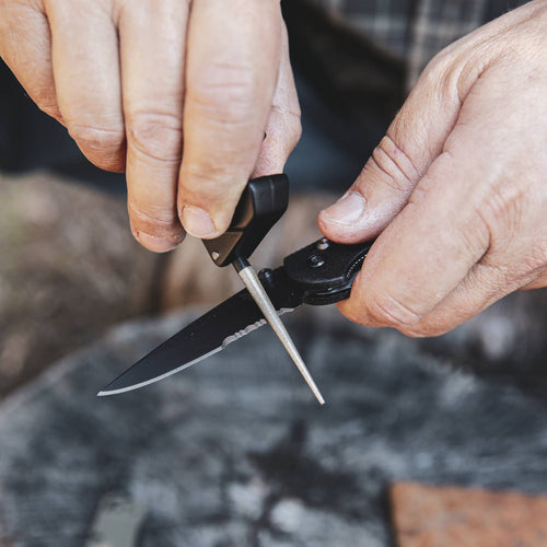 Person sharpening a serrated blade using a compact black and yellow handheld sharpening tool, shown outdoors, suited for Farm, Garden & Tool, Kitchen & Food, Sharpening, and Knives & Axes collections.