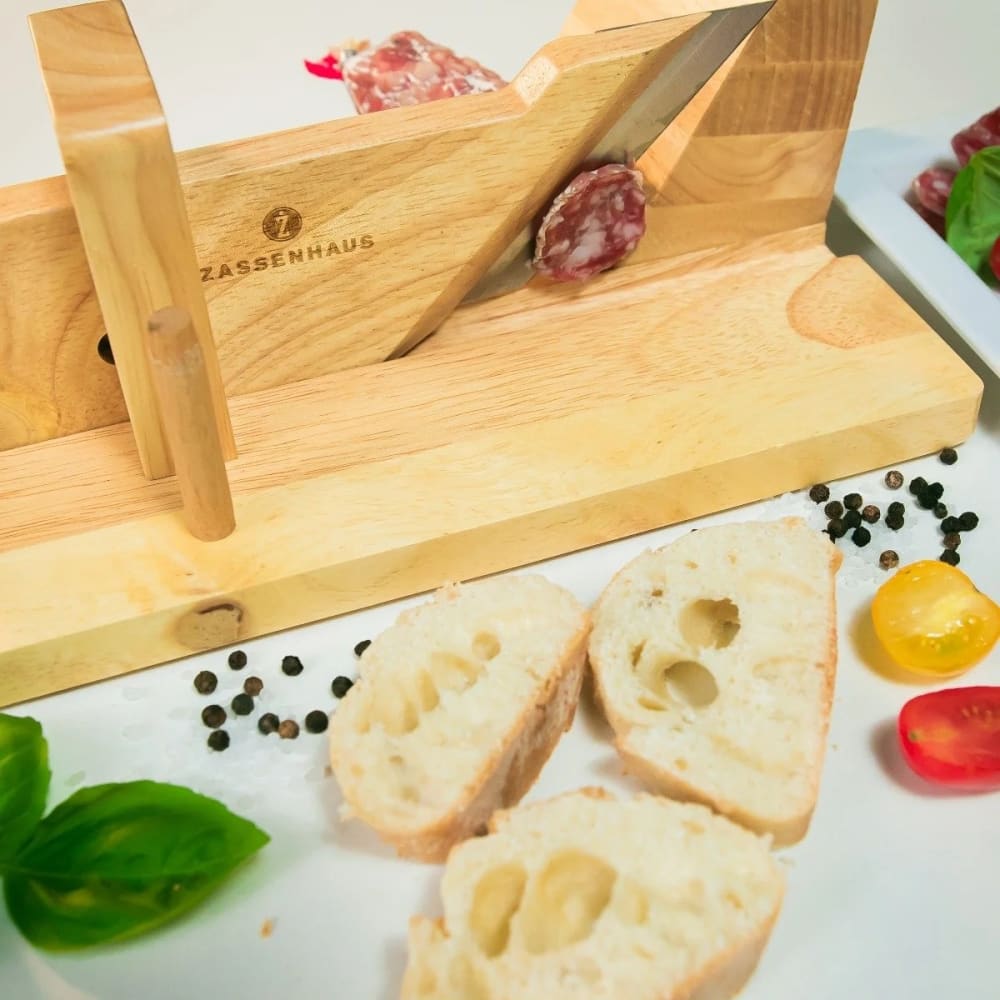 Wooden gourmet charcuterie slicer with sharp angled blade slicing cured sausage, surrounded by fresh bread slices, cherry tomatoes, basil, and peppercorns, ideal for Kitchen & Food and Food Prep collections.