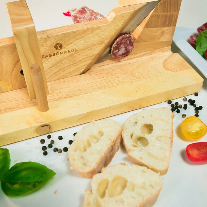 Wooden gourmet charcuterie slicer with sharp angled blade slicing cured sausage, surrounded by fresh bread slices, cherry tomatoes, basil, and peppercorns, ideal for Kitchen & Food and Food Prep collections.