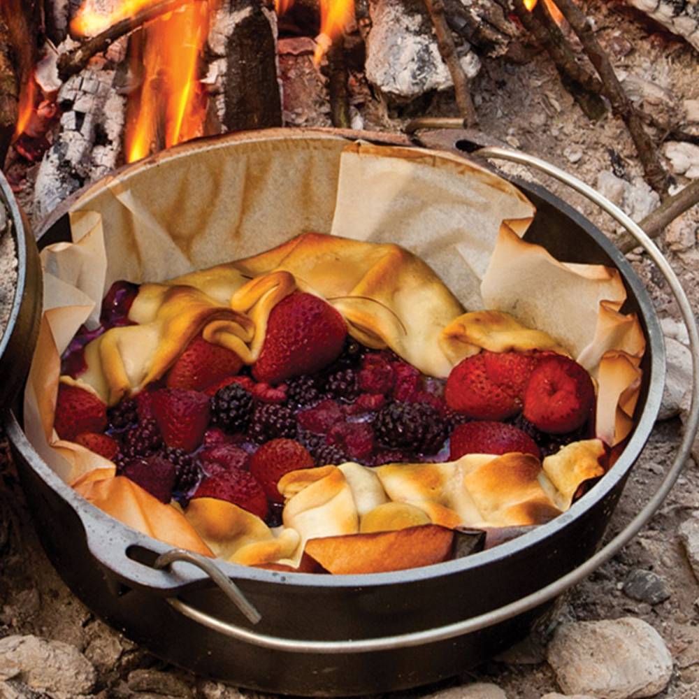 Handcrafted Lodge cast iron camp Dutch oven with built-in legs cooking a mixed berry cobbler outdoors over campfire coals, featured in Farm, Garden & Tool, Camping, Cast Iron, USA Made, Best Sellers, and Promo Eligible collections.