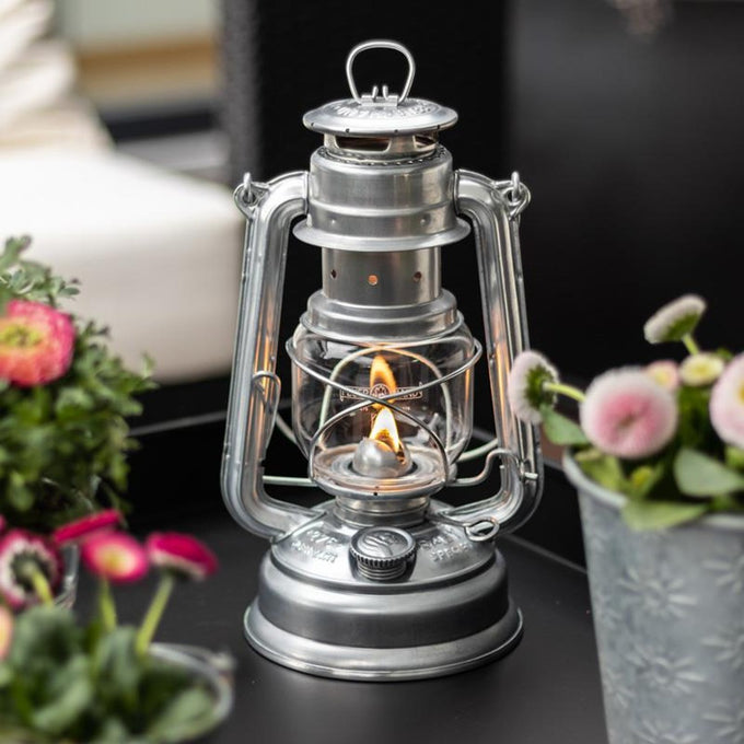 Unique handcrafted galvanized oil lantern with thermal heat-resistant SUPRAX globe, softly glowing indoors surrounded by vibrant floral arrangements, ideal for Camping, Lamps & Lighting, and Emergency Supplies collections.