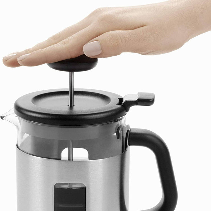 Hand pressing down the plunger on a stainless steel OXO Brew French Press Coffee Maker with black lid and handle, featured in Kitchen & Food, Brewing Coffee and Tea, Best Sellers, and Promo Eligible collections.