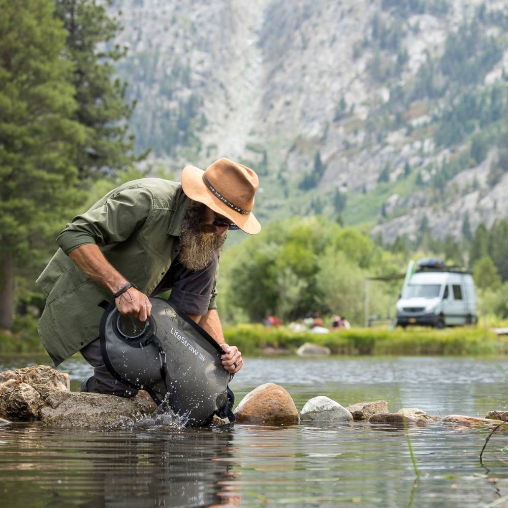 Man wearing a hat uses a black LifeStraw Peak Series gravity water filter to collect water from a mountain stream, surrounded by forest and rocky terrain, ideal for camping and emergency supplies.