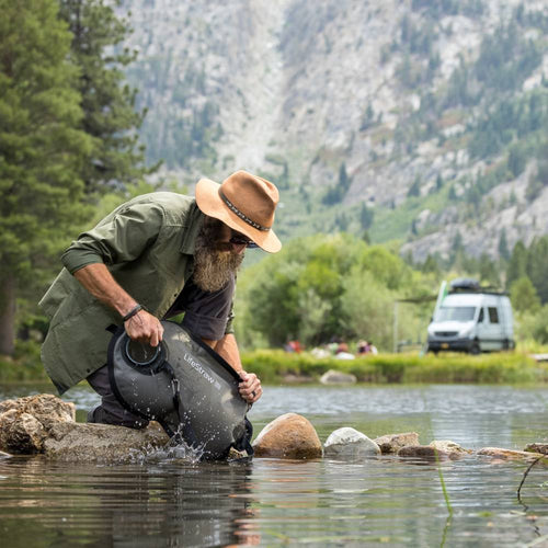 Man wearing a hat uses a black LifeStraw Peak Series gravity water filter to collect water from a mountain stream, surrounded by forest and rocky terrain, ideal for camping and emergency supplies.