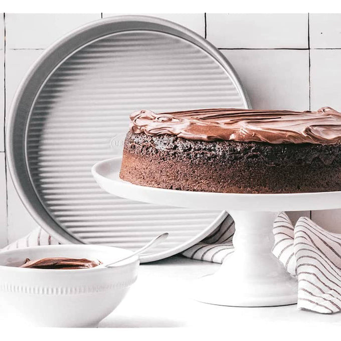 9-inch aluminized steel round cake pan with fluted surface for even baking, displayed with a frosted chocolate cake, white cake stand, and bowl of chocolate frosting, ideal for Kitchen & Food and USA Made collections.